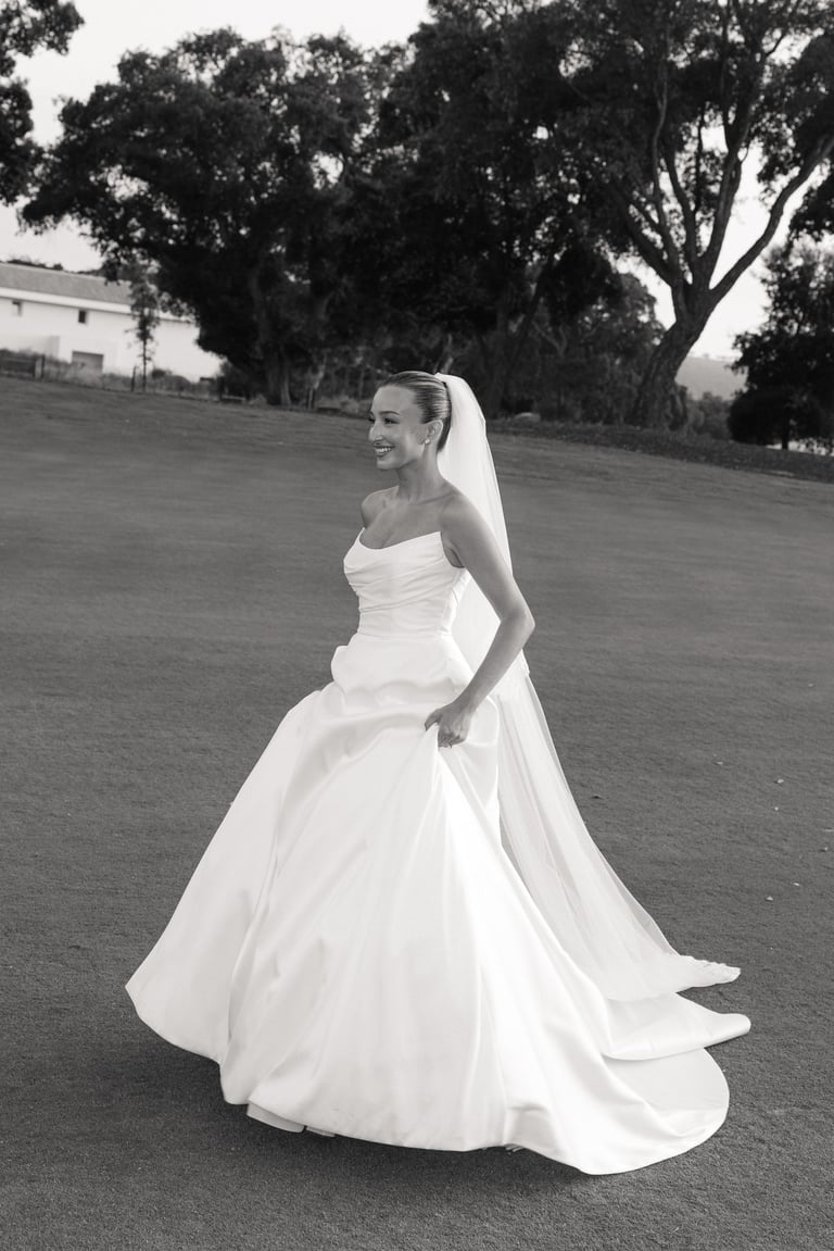 Black and white portrait of a bride in a white ball gown with veil standing on a grassy field with trees in the background