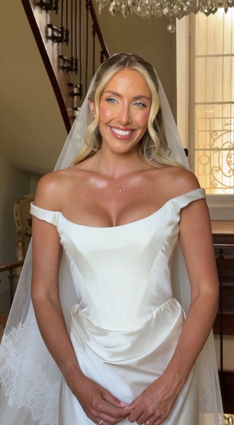 Woman in white off-shoulder wedding dress and veil smiling at camera in elegant room with chandelier and windows