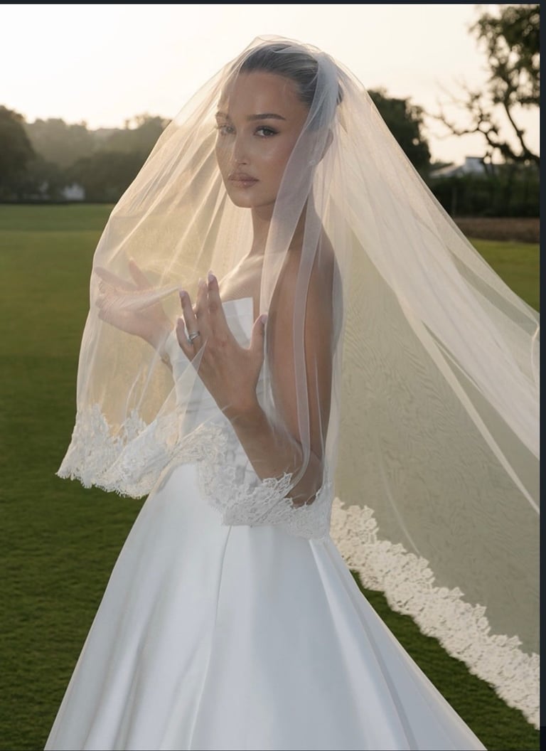 Bride in white wedding dress with flowing veil standing in outdoor garden setting at sunset
