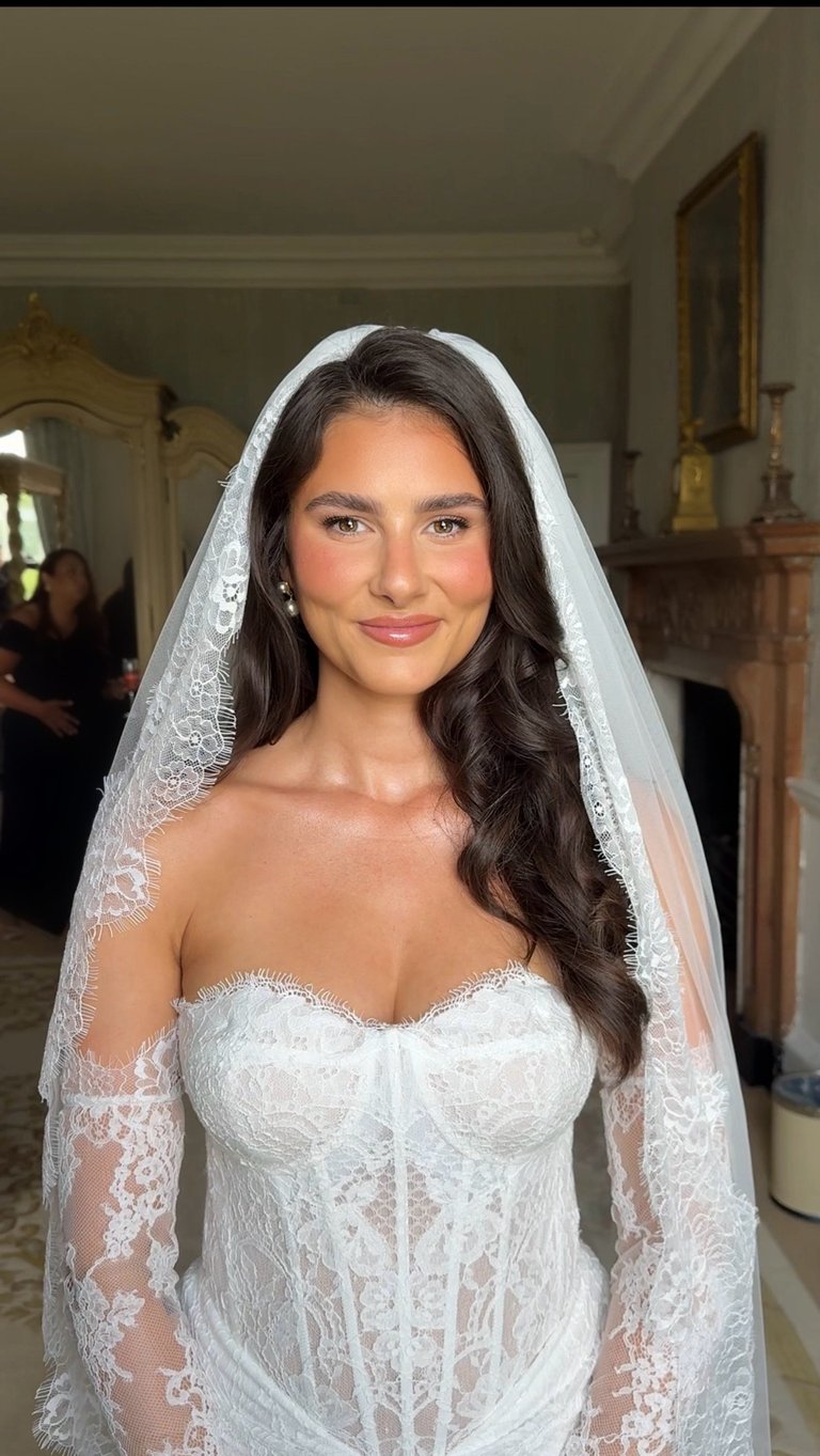 Young woman in white lace wedding dress and veil with long dark hair, smiling at camera in elegant interior setting