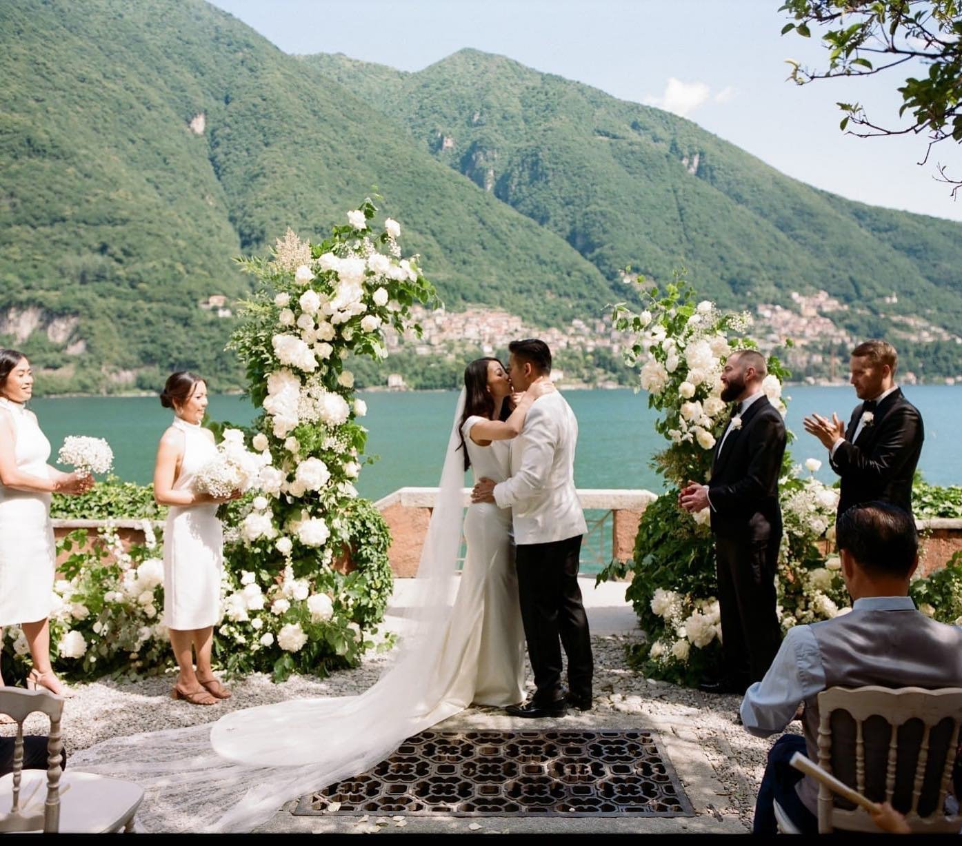 Bride and groom exchange vows at outdoor lakeside wedding ceremony surrounded by white floral arches and mountains