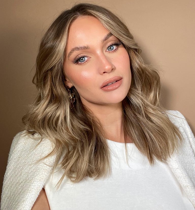 Woman with long wavy light brown hair, blue eyes, and neutral makeup wearing a white knit sweater against a beige background
