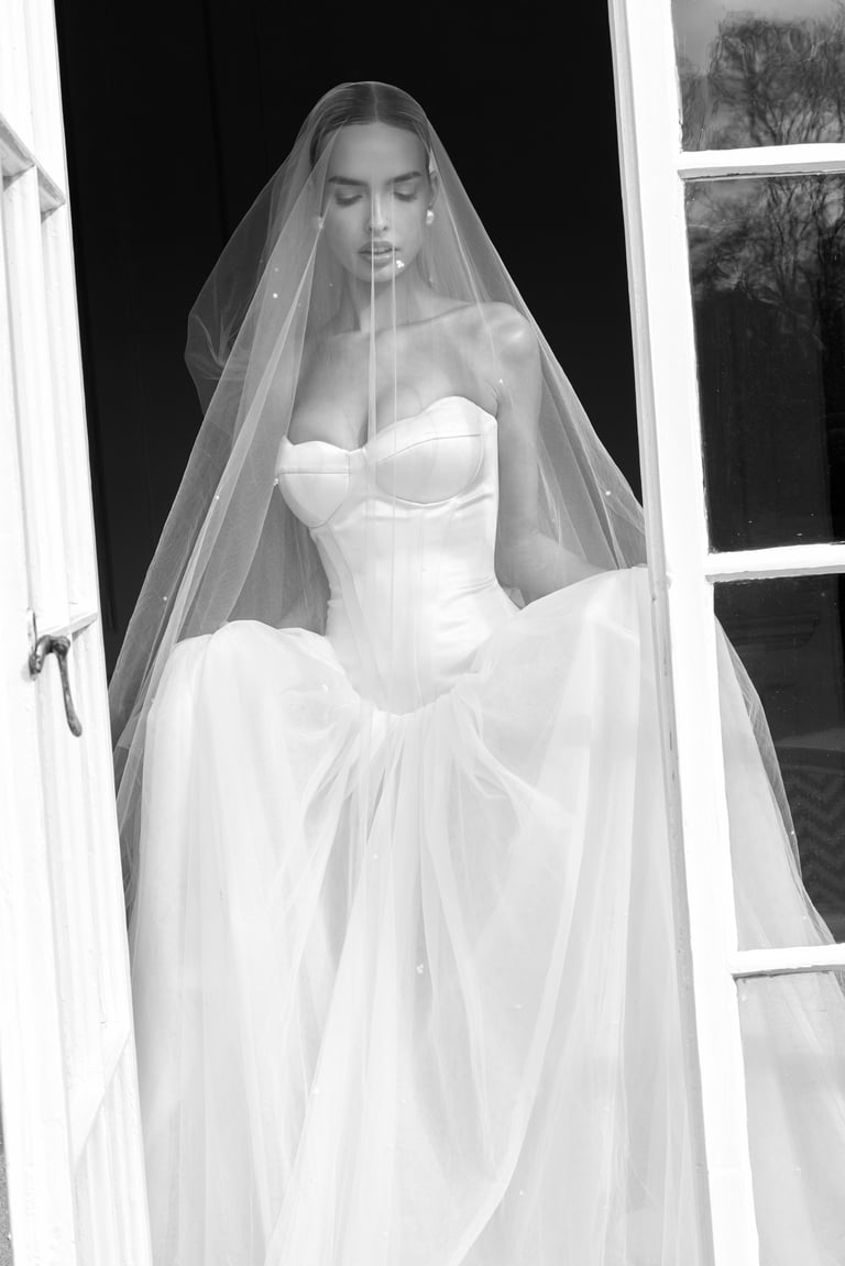 Woman in white off-shoulder wedding gown with flowing veil, standing in black and white portrait photo