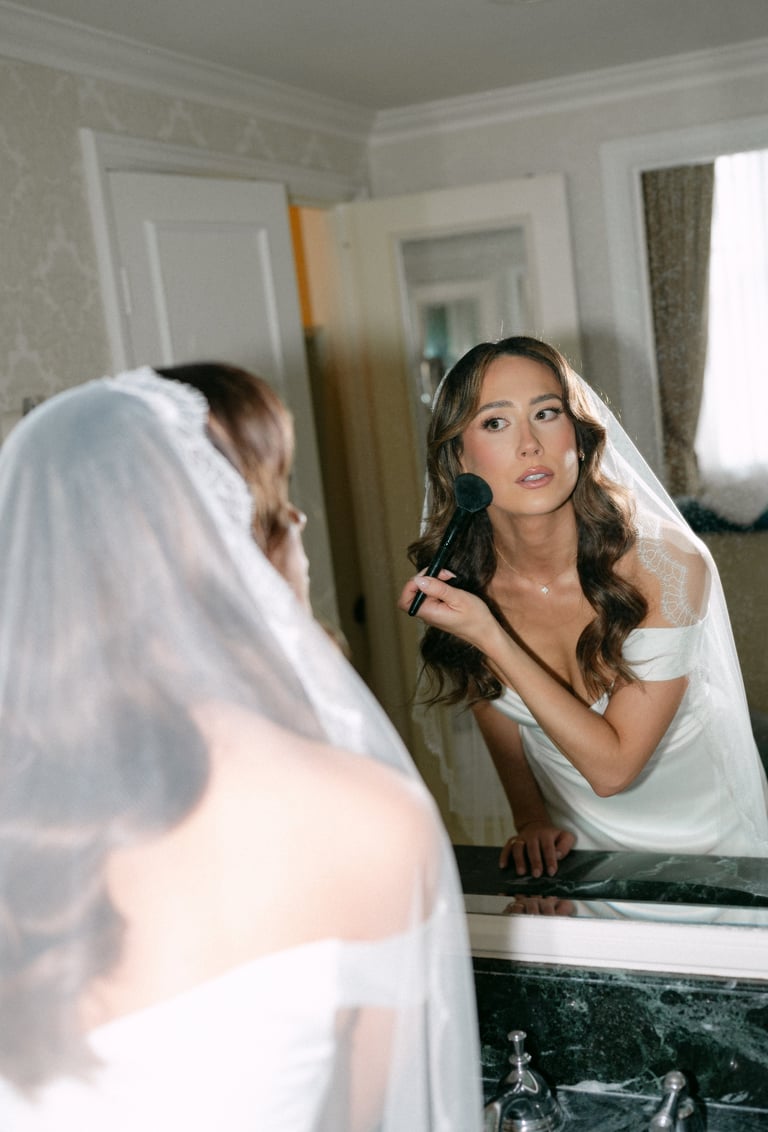 Bride in white veil applying makeup with brush in bathroom mirror, preparing for wedding