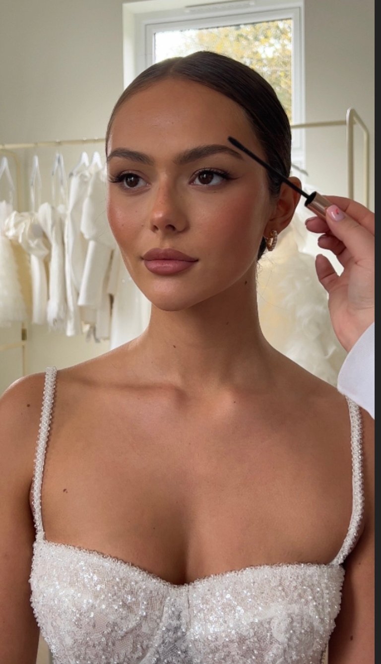 Woman getting eyebrow makeup applied in a bathroom, wearing a white beaded top