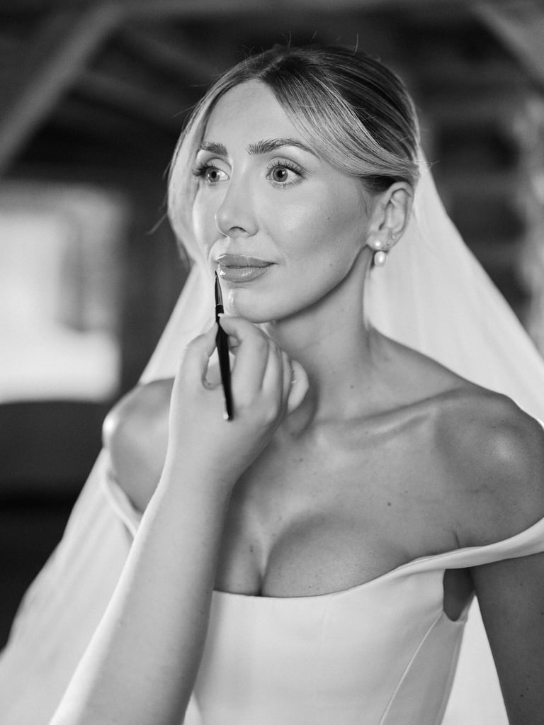 Black and white portrait of a woman holding a makeup brush to her face, wearing an off-shoulder white top and veil