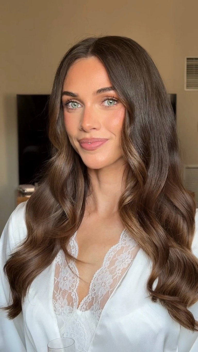 Woman with long brown wavy hair wearing white lace top, posed indoors against beige wall