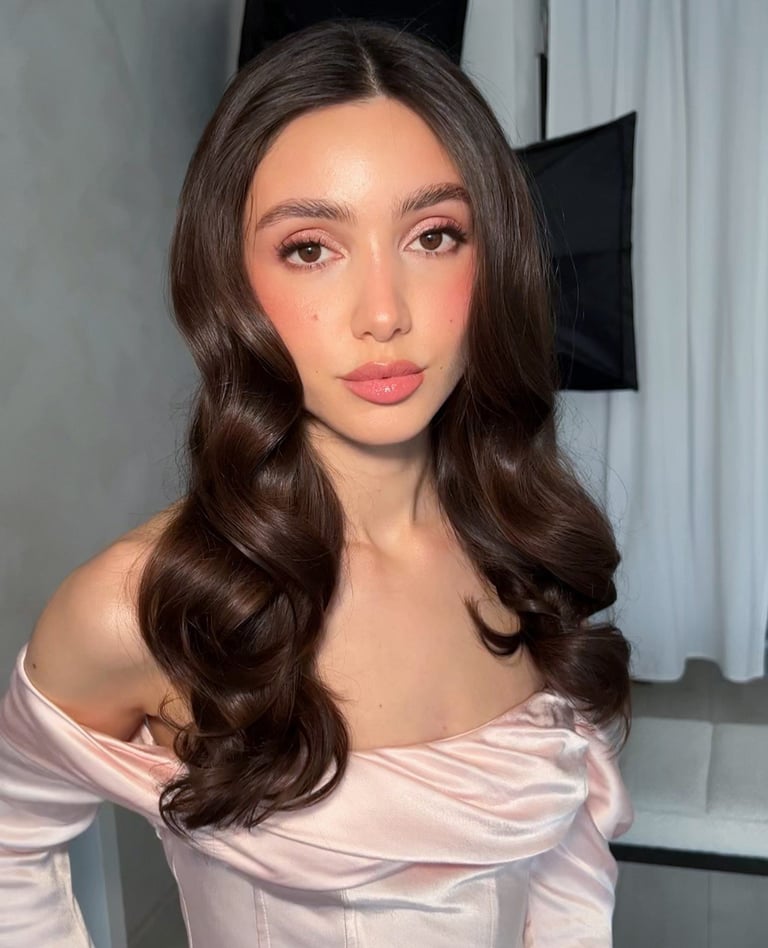 Woman with long wavy brunette hair and pink lipstick wearing an off-shoulder pink satin blouse against a white and black background