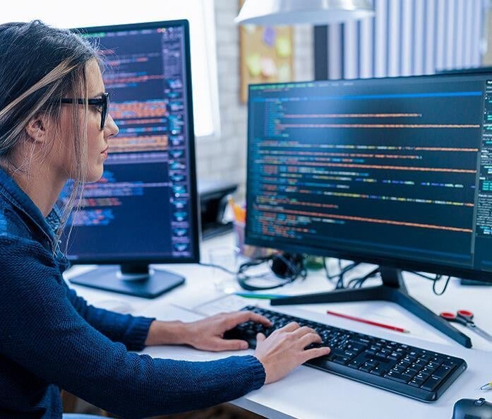 Developer working at a desk with dual monitors displaying code, wearing glasses and typing on a keyboard