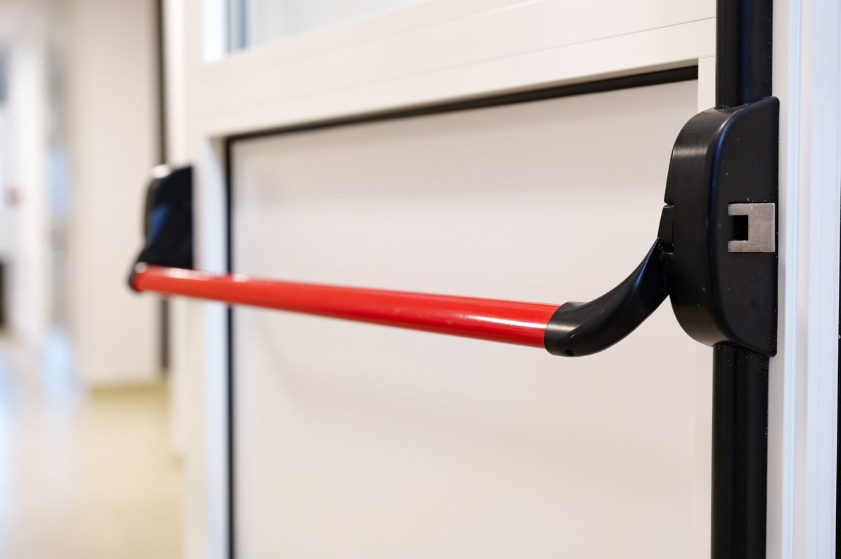 Black and red panic bar exit device mounted on white door frame in hallway