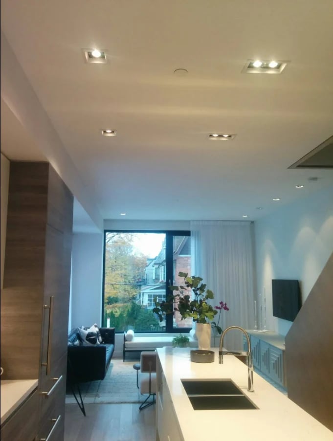 Modern kitchen with white countertops and sink, opening to a bright living room with large window and natural light