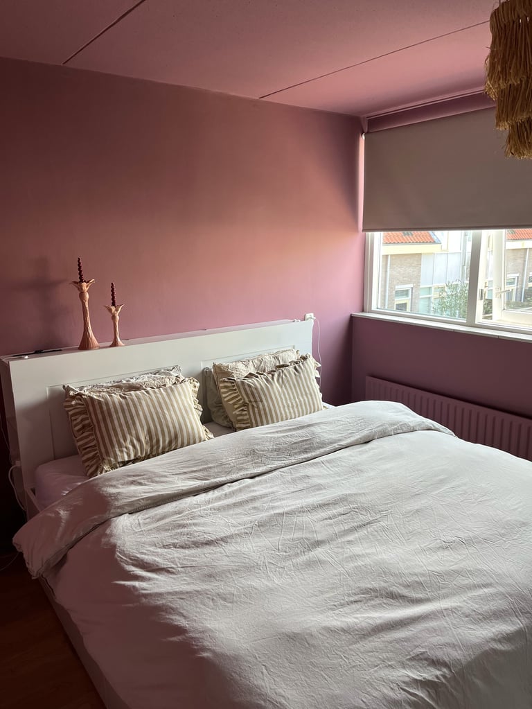 Moderne slaapkamer met roze muren, wit bed met gestreepte kussens, raam met grijze jaloezieën en decoratieve kaarsen op nachtkastje