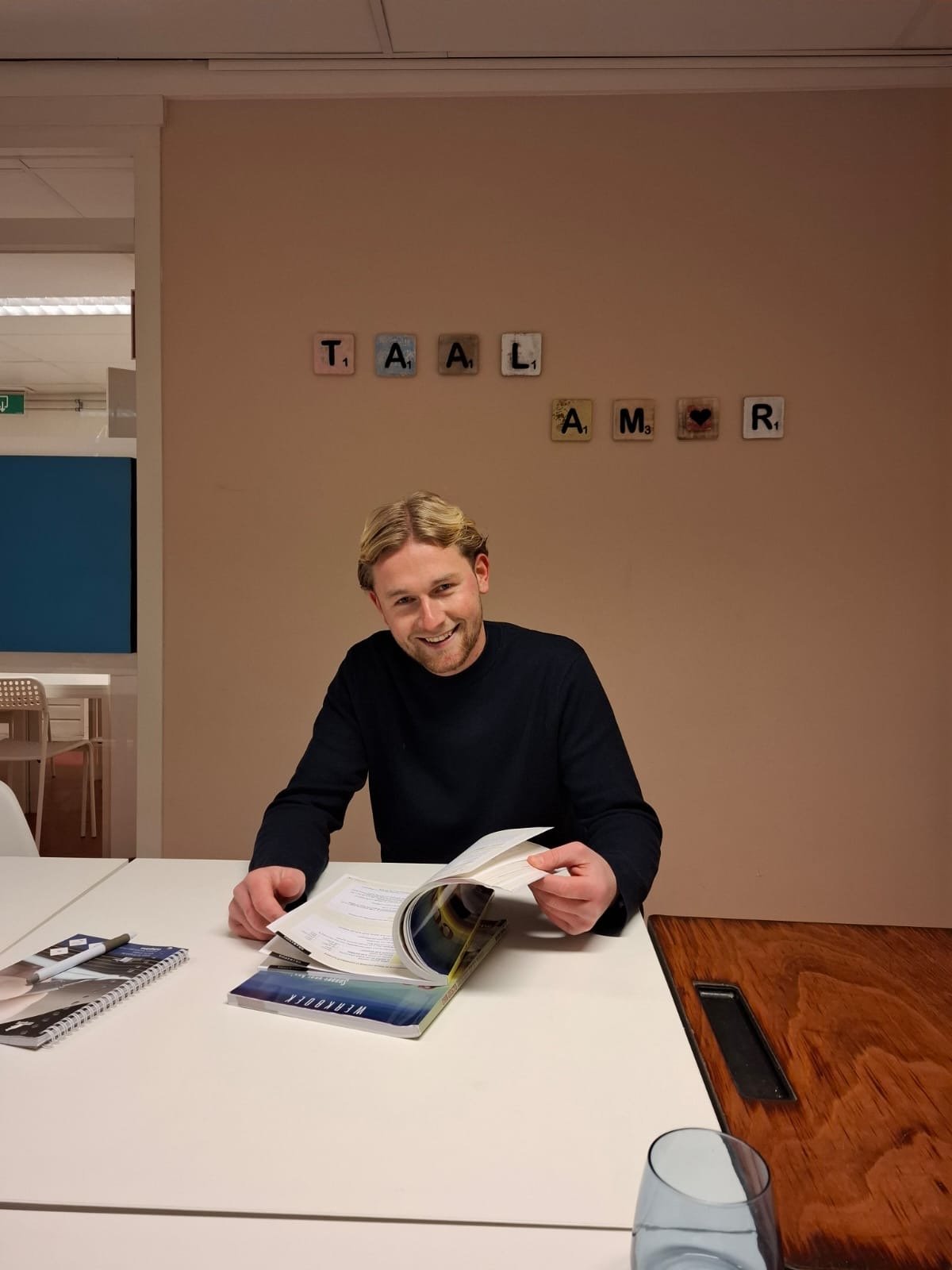 Man in zwarte trui lachend tijdens lezen van tijdschrift aan tafel onder TAAL AMR bord aan muur
