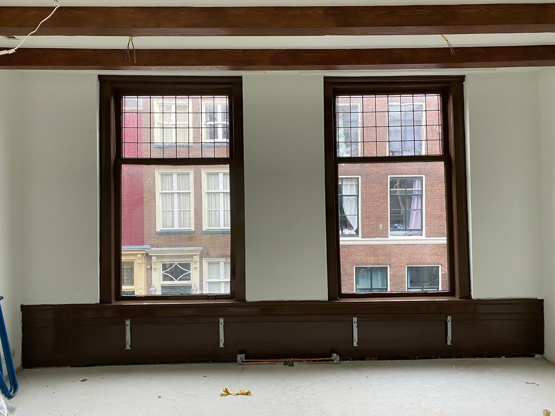 Twee grote donker omlijste ramen in lege kamer met lichtgroene muren en zichtbare plafondbalk richting stadsgebouwen