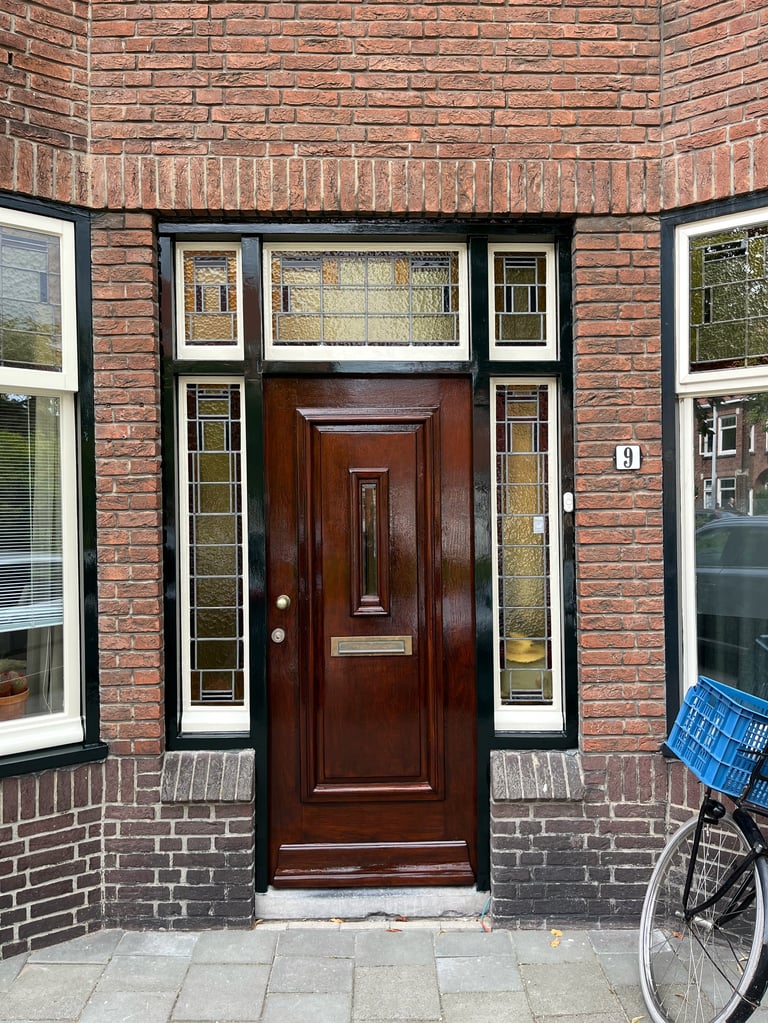 Rode bakstenen gevel met donkere houten voordeur tussen twee ramen, fiets ernaast