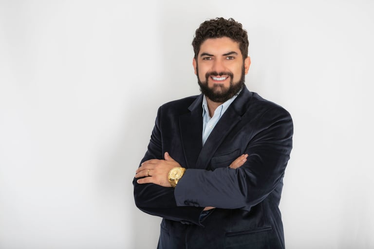 Professional man with dark beard wearing navy blazer and light blue shirt, smiling with arms crossed against white background