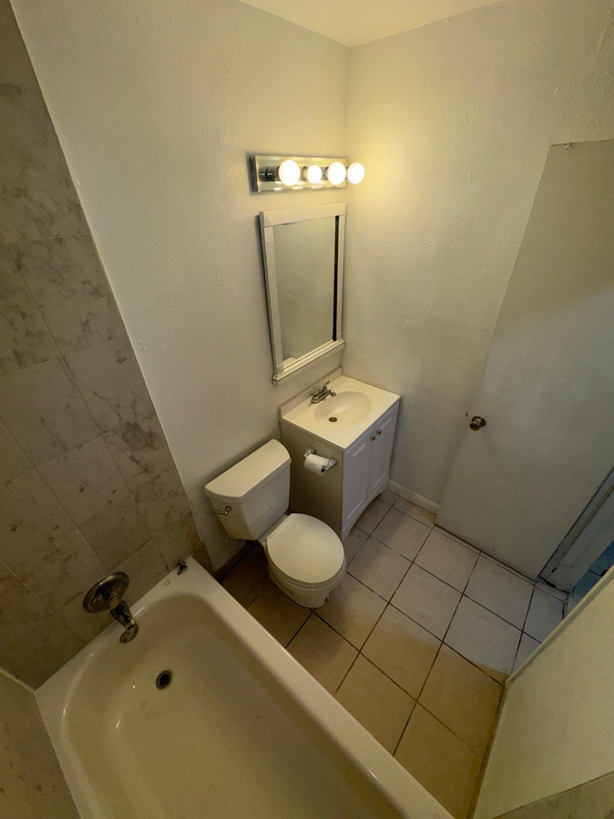 Small bathroom with toilet, pedestal sink, mirror, and vanity light fixture above, tile flooring and walls