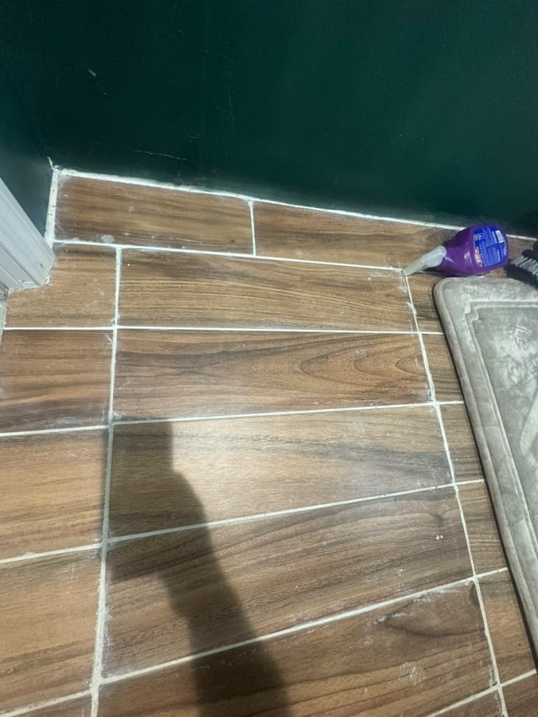 Wood-grain tiles with white grout lines being installed on a floor, with a purple adhesive bottle visible in the corner