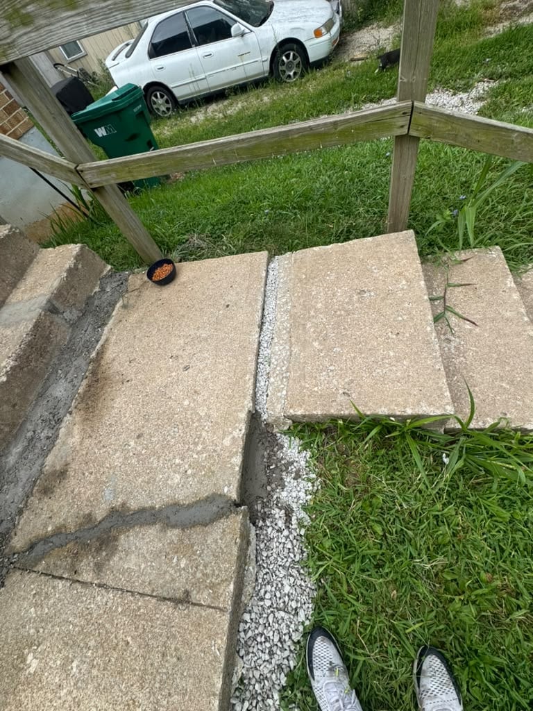 Overhead view of a concrete sidewalk with grass growing between cracks, white car parked in background driveway
