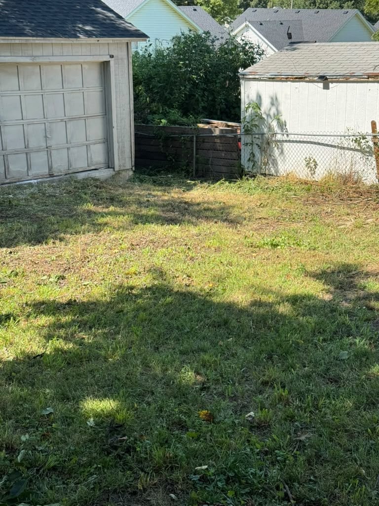 Suburban backyard with overgrown grass, white garage, wooden fence, and residential houses in background