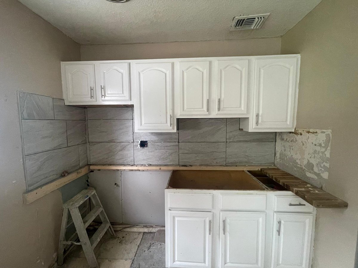 Kitchen renovation in progress showing white cabinets, exposed wood countertops, and tile backsplash with construction materials