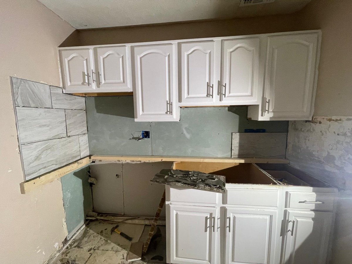 Kitchen under renovation with white cabinets, exposed walls, and construction materials scattered throughout the space