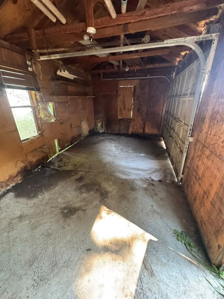 Interior of an old, deteriorating wooden garage with concrete floor, sunlight streaming through windows, and weathered wooden walls and doors
