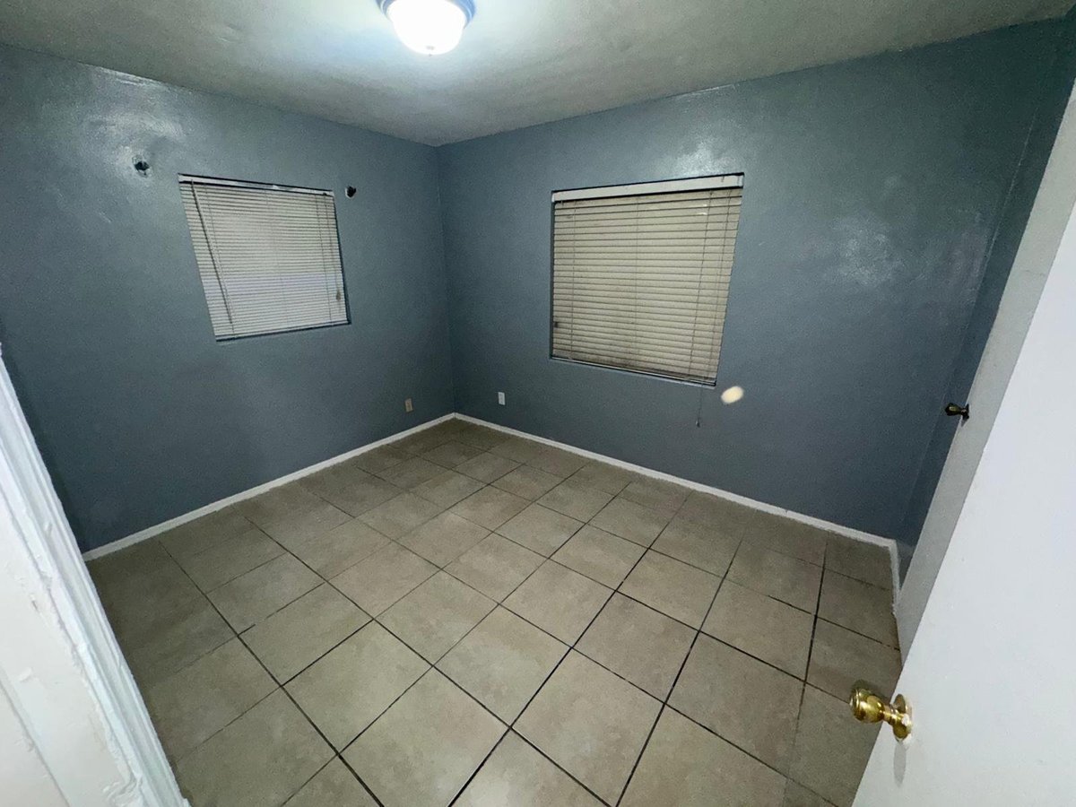 Empty room with teal walls, tile flooring, two windows with blinds, and a doorway with brass handle