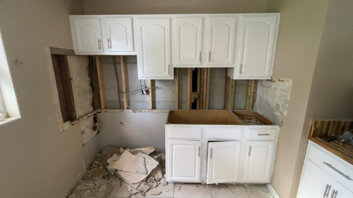 Kitchen under renovation with white cabinets removed, exposing wall framing and debris on floor