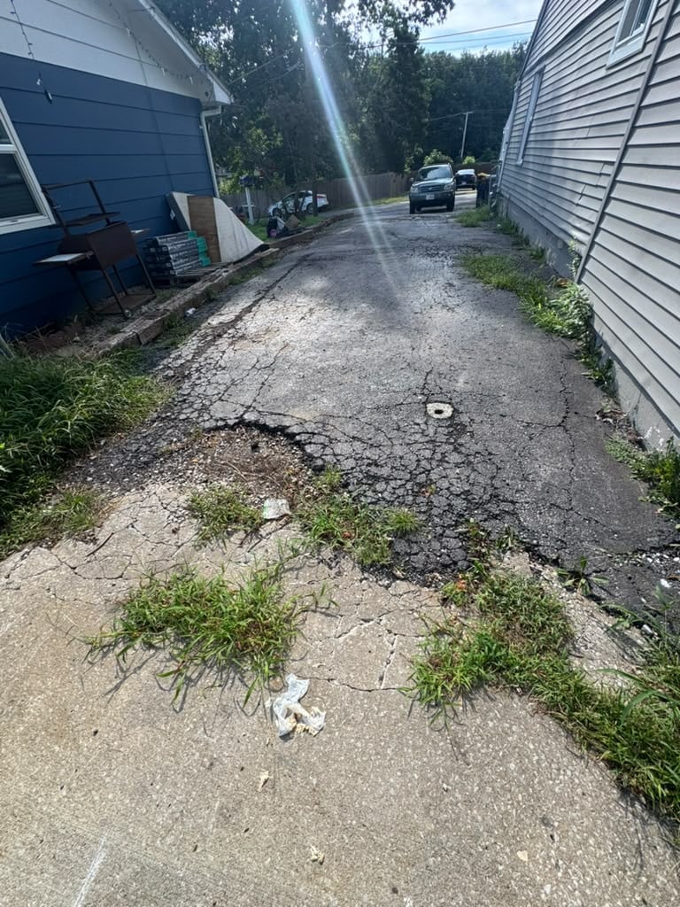 Narrow gravel driveway between two residential houses with overgrown weeds and parked vehicles in background