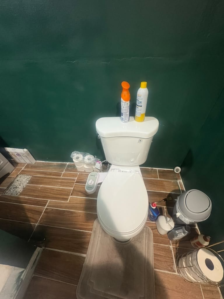 White toilet in a bathroom with various personal care products and supplies arranged on and around it against a dark teal wall