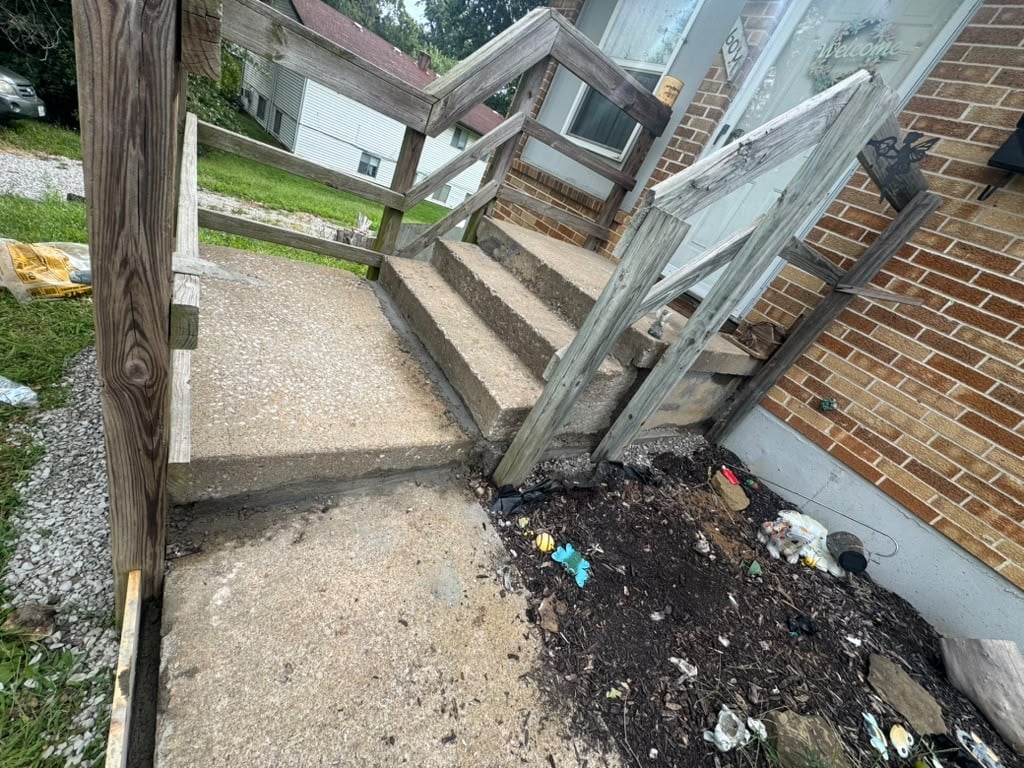 Outdoor pathway alongside brick building with wooden gate, concrete steps, and scattered debris on ground