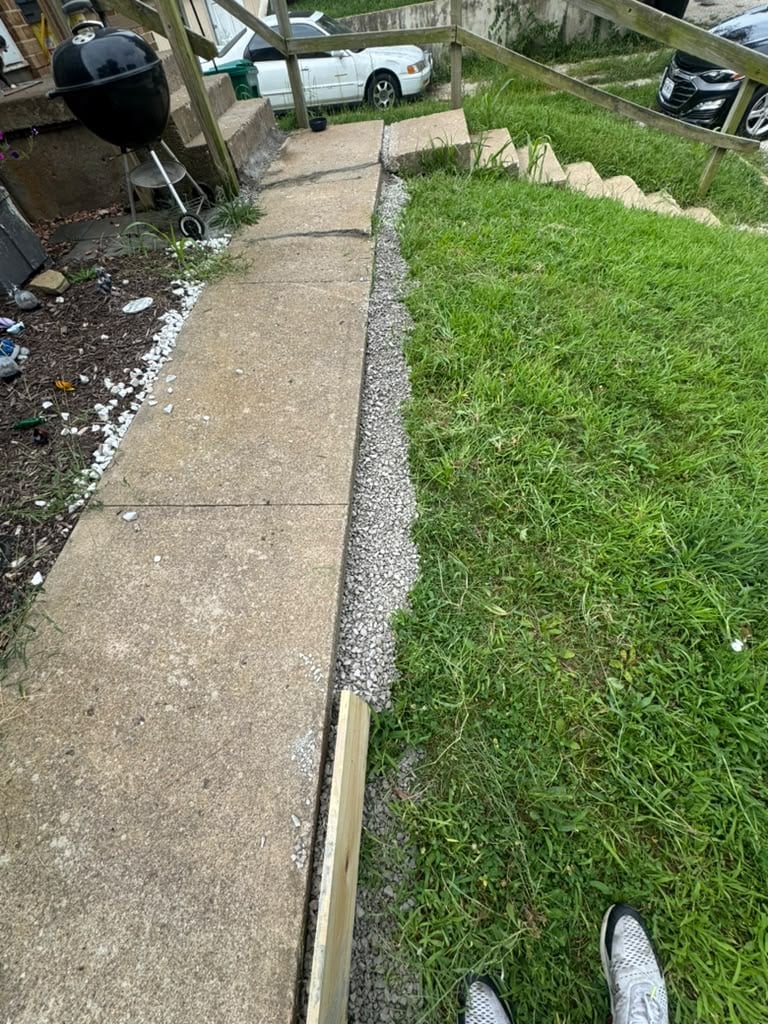 Concrete sidewalk with gravel border separating from green lawn, viewed from ground level with parked cars visible in background