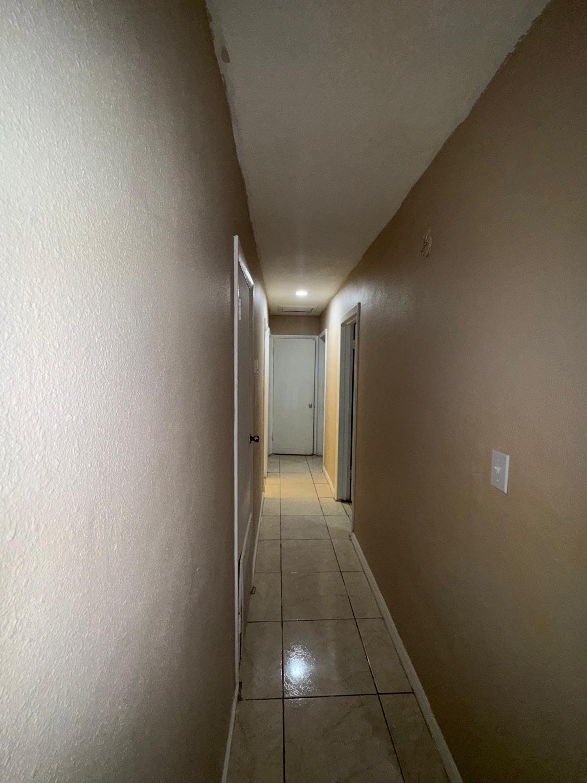 Long narrow hallway with beige walls, tiled flooring, and doors on both sides leading to other rooms