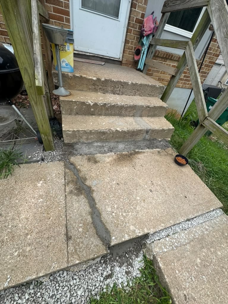 Weathered concrete patio and steps leading to a brick house with wooden fence and garden items nearby