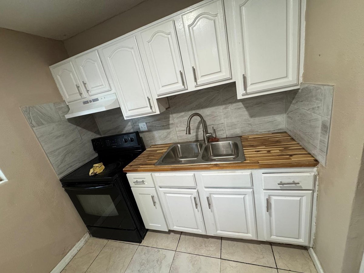 Small kitchen with white cabinets, stainless steel sink, black stove, wooden countertop, and gray tile backsplash