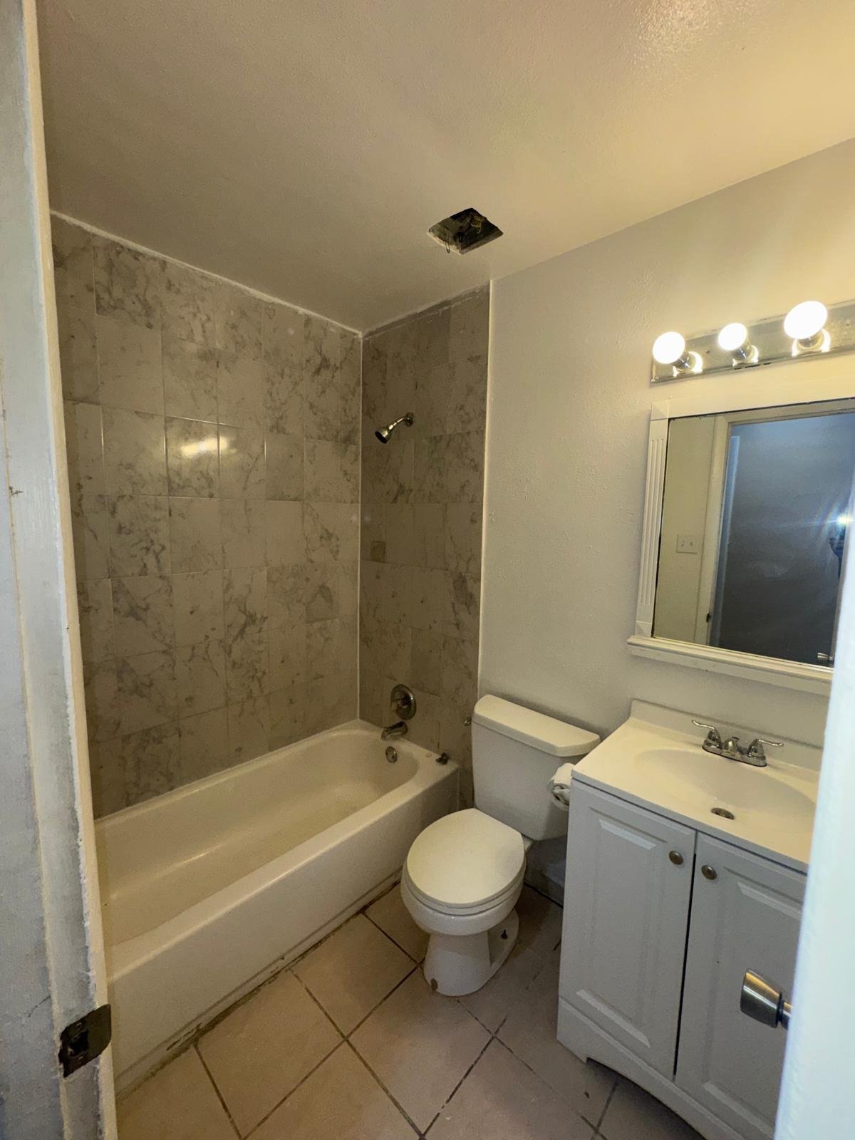 Modern bathroom with white vanity, mirror with lights, toilet, and bathtub with marble tile surround