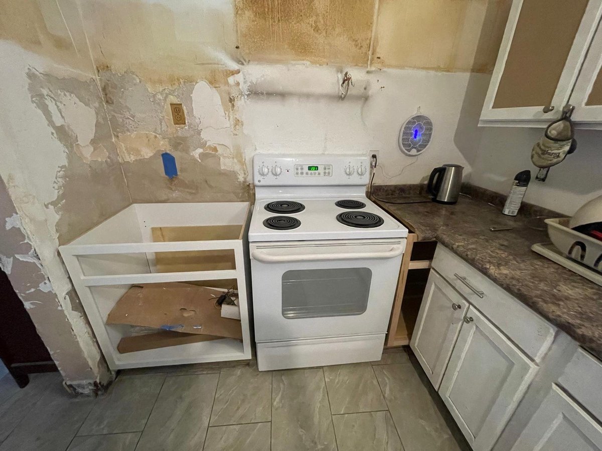 Kitchen renovation in progress showing white stove, drywall repairs, exposed cabinetry, and construction materials