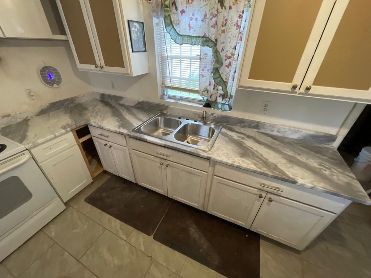 Kitchen with white cabinets, marble-look countertop, stainless steel sink, window above sink, and brown tile flooring