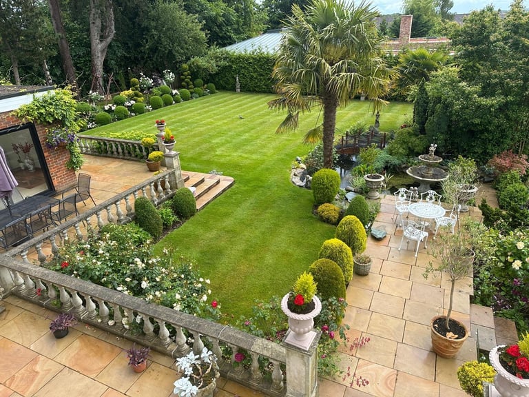 Aerial view of a large manicured garden with rolling lawn, palm tree, stone patio, and flowering plants along the borders
