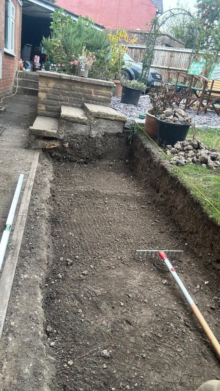 Excavated garden bed with soil and a measuring pole, surrounded by stone steps and planter boxes in a residential backyard