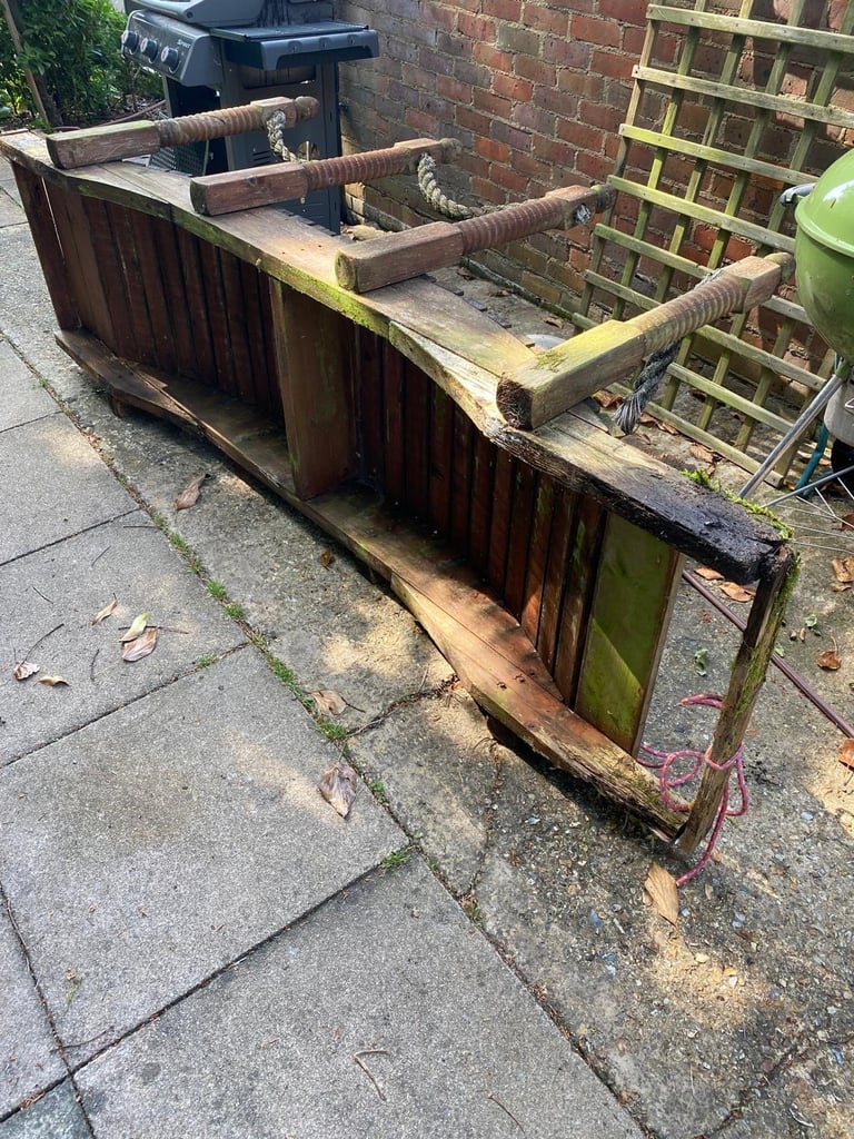 Weathered wooden garden trough with moss and algae buildup, sitting on a paved outdoor area near a brick wall