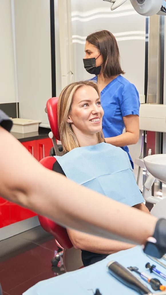 Smiling patient in dental chair with protective bib while masked dentist stands behind her in a modern dental office