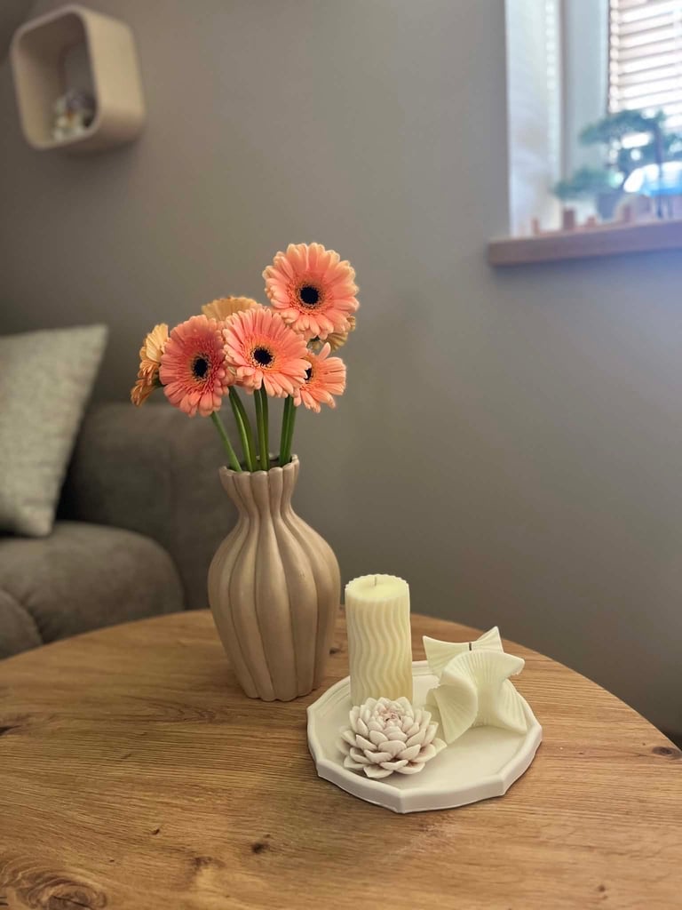 Coral gerbera daisies in a ribbed beige vase on a wooden table with decorative white items