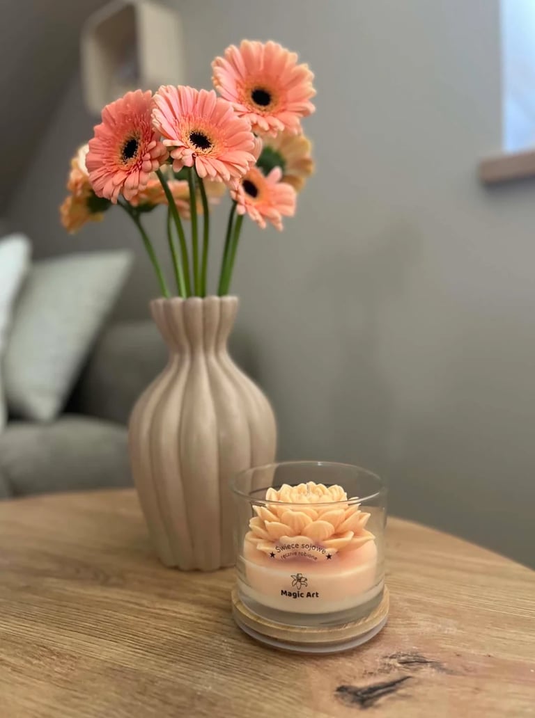Pink gerbera daisies in a white vase with a decorative candle on a wooden table in a modern living room