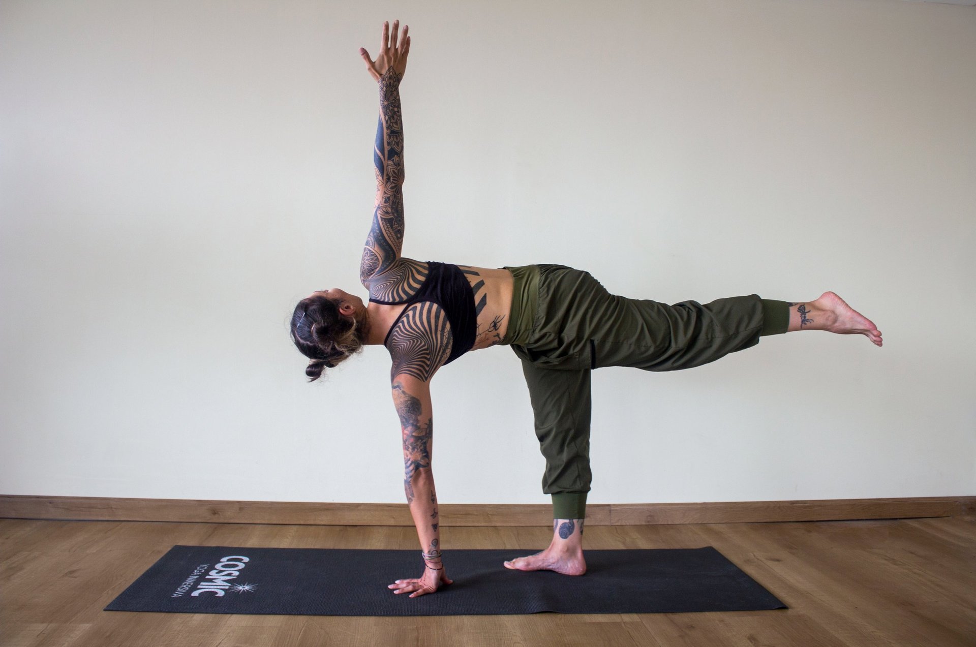 Tattooed man performing a one-armed yoga balance pose on a dark mat against a white wall
