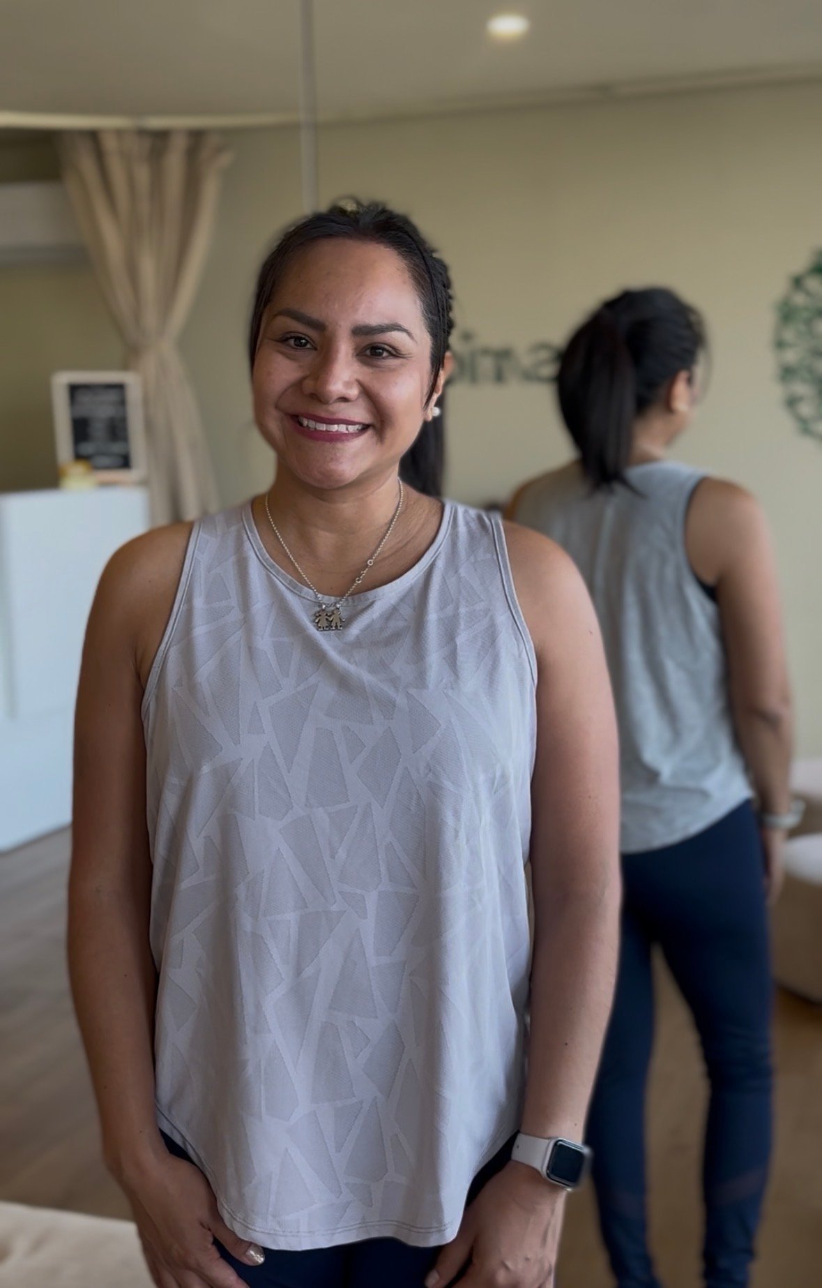 Woman smiling at camera in light gray sleeveless top with smartwatch, indoors with another person visible in background