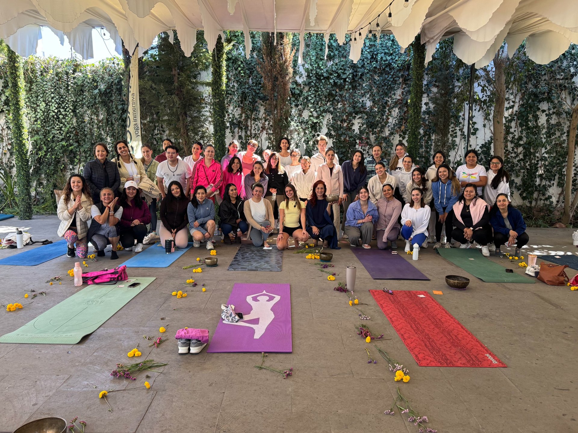Group of women posing under a draped tent with yoga mats on ground, surrounded by ivy-covered walls and decorative flowers