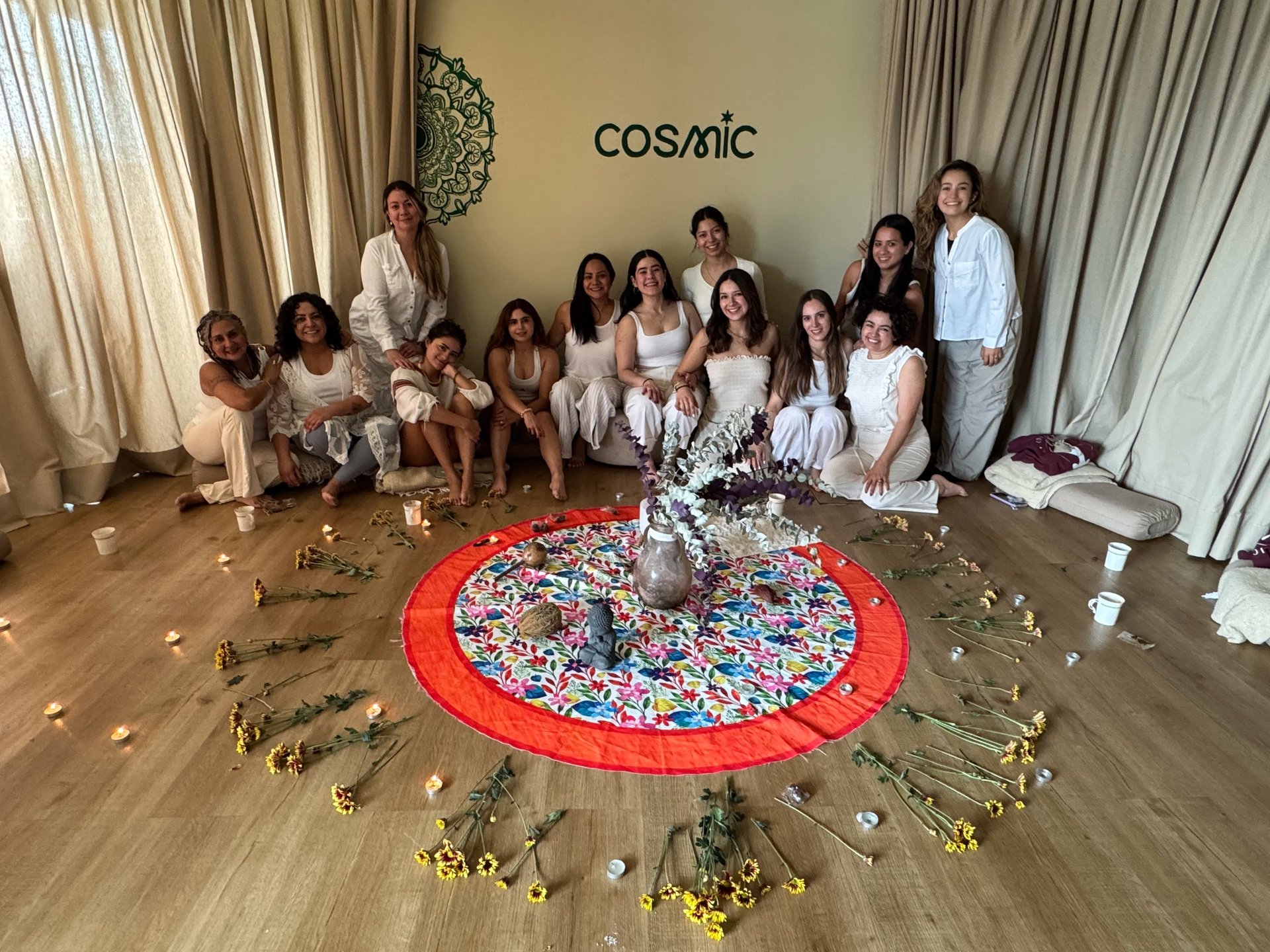 Group of people in white clothing standing around a red circular mandala art installation with gold accents on a wooden floor