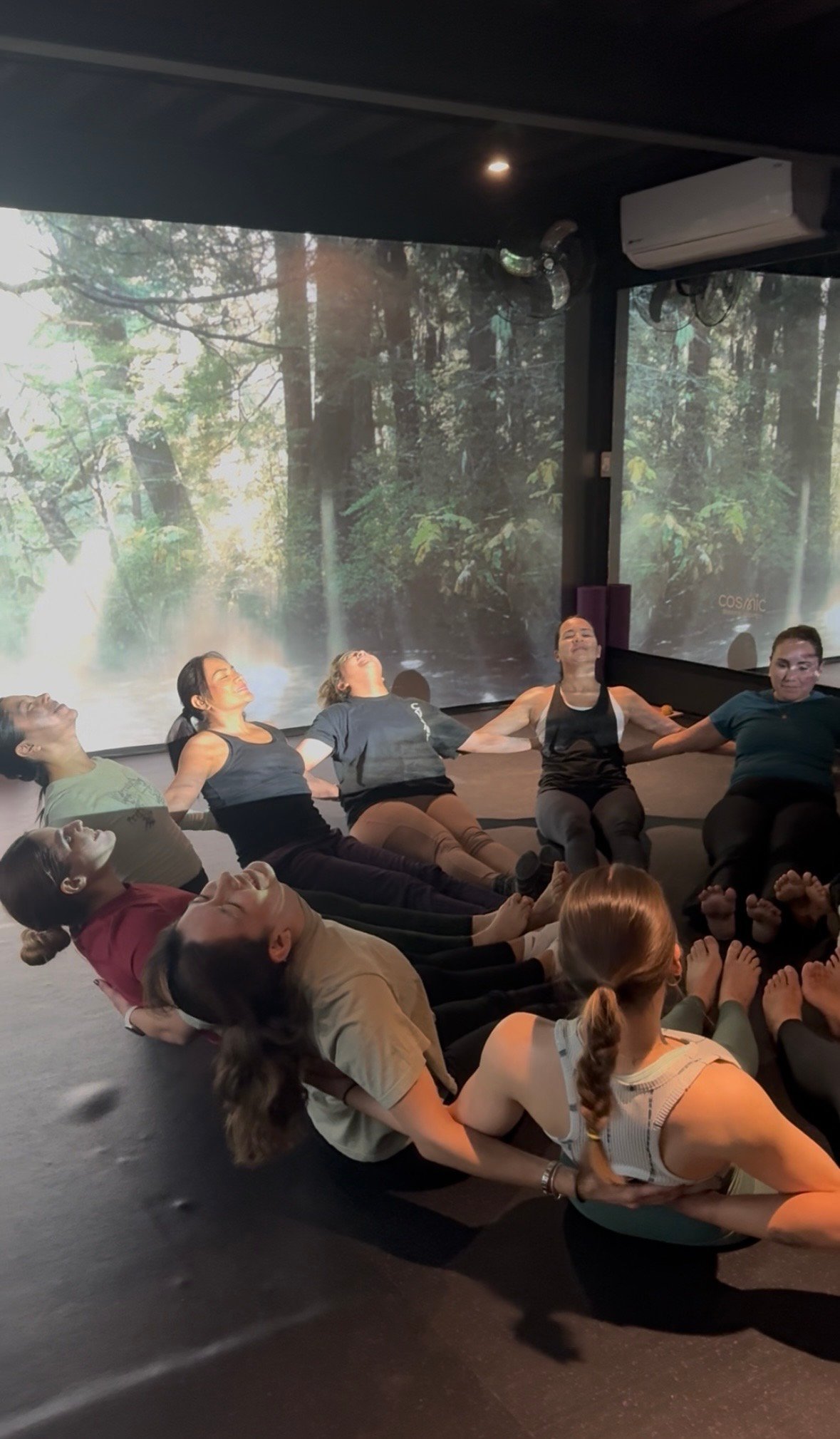 Group of people sitting on floor in circle during fitness class with forest scenery projected on large screens behind them